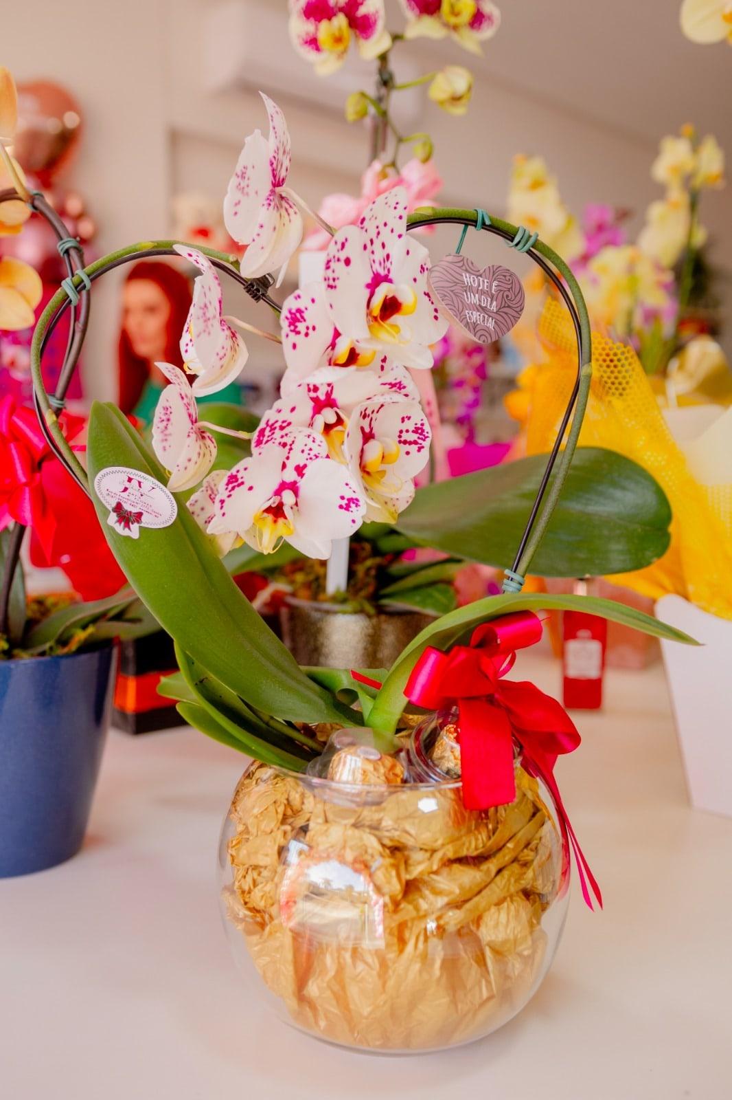 Orquídea branca e rosa num vaso de vidro com bombons e laço vermelho, do jeito que chega na Assinatura Floral